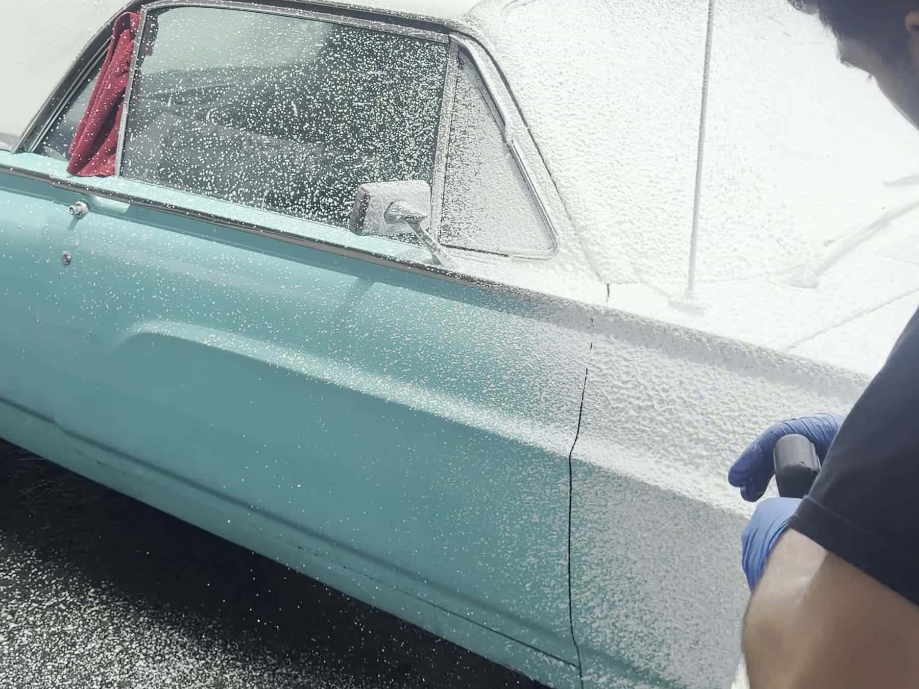 A classic Ford Thunderbird covered in snow foam, being washed by hand